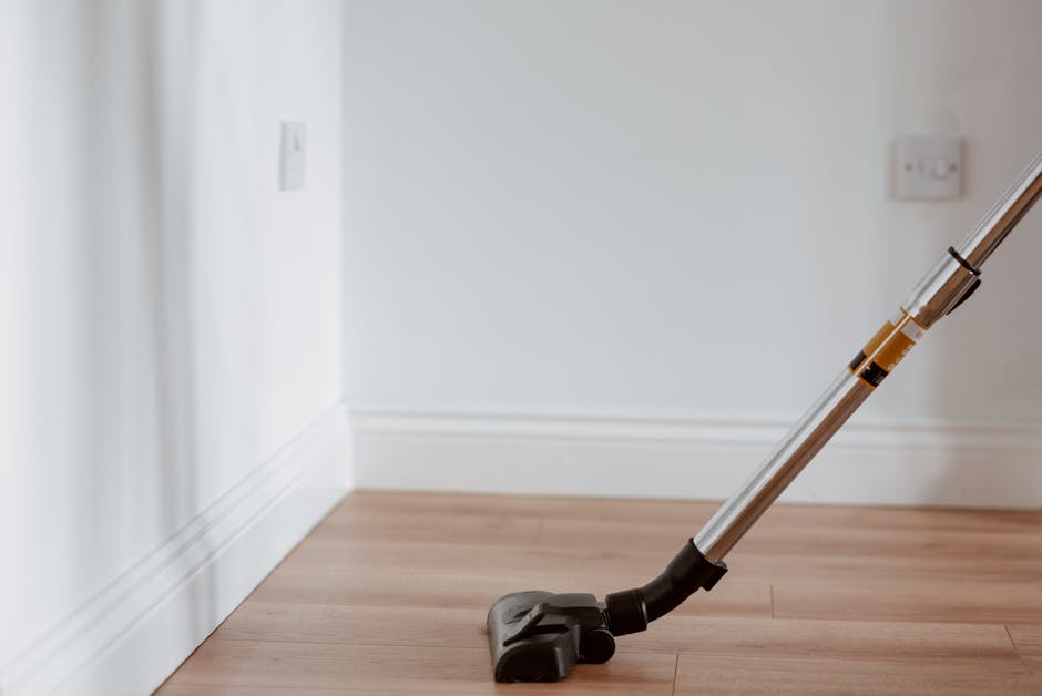 A professional cleaner wearing a black uniform and black gloves is preparing to disinfect a wooden dining table in a bright, modern bedroom. The table, made of polished dark wood, is situated near a w