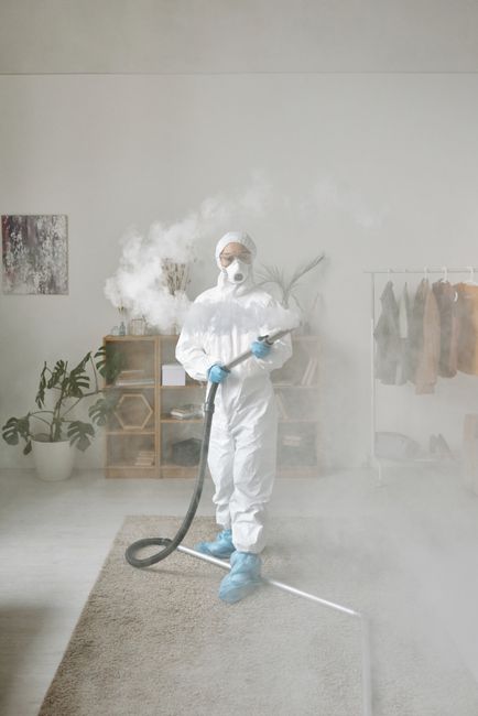 A professional cleaner dressed in white protective gear, including a hood, mask, gloves, and shoe covers, is performing surface cleaning on a cream-colored carpet in a modern, well-lit living room. Th