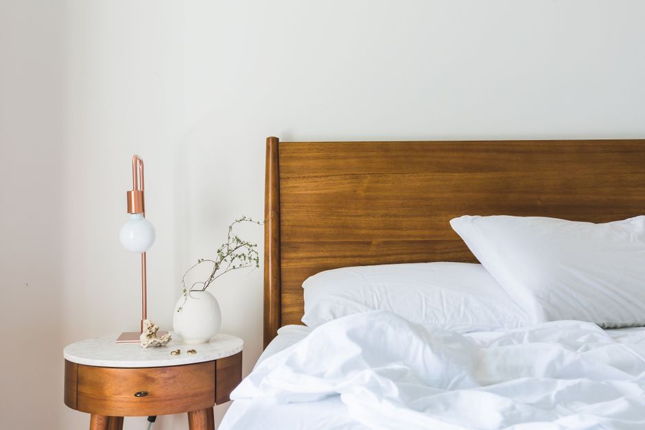 A neatly made bedroom featuring a large, upholstered blue bed with a freshly cleaned white mattress. The room has a polished wooden floor and is illuminated by natural light from a window covered with