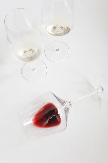 A hand holding a wine glass tilted upside down with red wine spilling out, creating a splash on a plain white background. The glass is clear and polished, and the spill appears fresh with liquid flowi